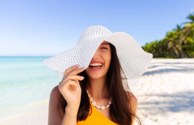 Sahilde arka planda açık mavi okyanusla duran hasır şapkalı genç kadın portresi, kameraya gülümseyen kadın, kumlu sahilde yürüyen kadın.