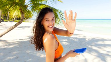 Gülümseyen kadın elinde güneş kremi şişesi tutarken ve parmağında krem lekesi gösterirken, arka planda palmiyeler ve okyanusla sahilde dururken, panorama