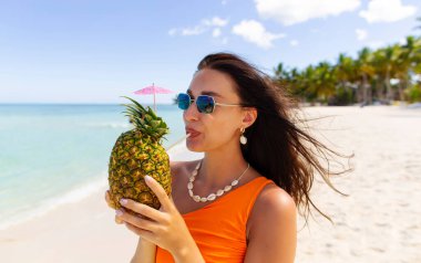Elinde samanla ananas içeceği tutan, kumlu tropik ada sahilinde yürürken taze meyve suyu ya da kokteyl içen bir kadın.