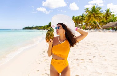 Mayo ve hasır şapkalı kadın taze ananas tutuyor ve taze meyve suyu içiyor, okyanus kıyısında duruyor, mükemmel cennet manzarasının tadını çıkarıyor.