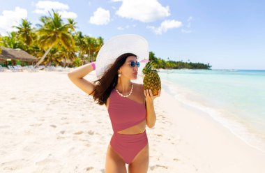 Rahat bir kadın tropik adanın okyanus kıyısında yürüyor, ananas tutuyor ve meyve suyu veya kokteyl içiyor, bedava uzay.