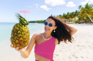 Cennet tatili. Pembe mayo giyen mutlu bayan ananaslı kokteyl tutuyor, kumlu tropik plajlarda yürüyor ve iyi günlerin tadını çıkarıyor.