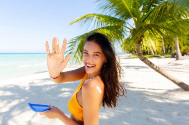 Güneş kremi şişesini tutan genç ve mutlu bir bayan. Plajda durup kameraya gülümsüyor.