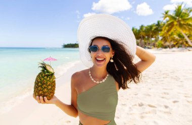 Güzel bir bayanın portresi taze ananas tutuyor ve tropik tatilinin tadını çıkarıyor, kumsalda yürüyor ve kameraya gülümsüyor.