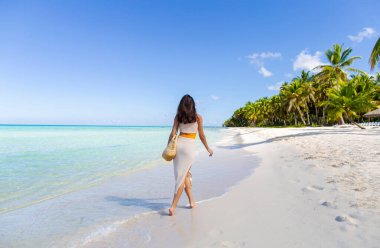 Tropikal adanın kumlu sahilinde örülmüş tunik giymiş genç bir kadının arka görüntüsü.