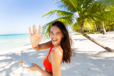 Güzel genç bir kadın vücuduna güneş kremi sürüyor ve parmağında krem lekesi gösteriyor. Cennetin tadını çıkarıyor.