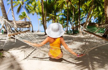 Hasır şapkalı, tanınmayan genç bir kadın cennetteki bir plajda, palmiye ağaçlarının altında oturuyor.