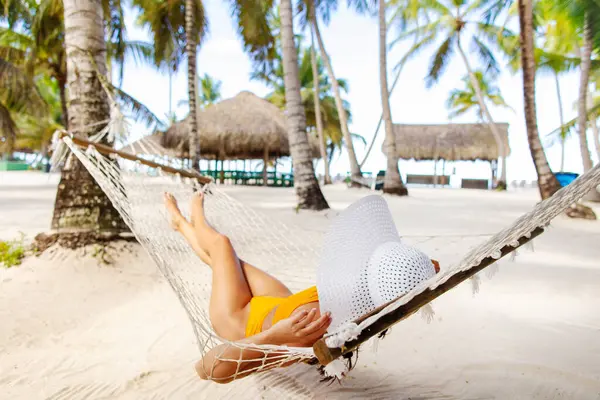 Hasır şapkalı bir kadın okyanus kıyısındaki palmiye ağaçlarının altında uzanmış tropikal tatilinin tadını çıkarıyor.