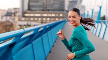 Heyecanlı zinde bir kadın şehir köprüsünde tek başına koşuyor ve kameraya gülümsüyor, güzel bir manzarayla dışarıda çalışmanın tadını çıkarıyor.