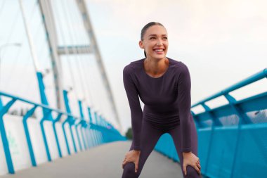 Heyecanlı atletik kadın idman yaparken ve koşarken nefes alıyor, şehirde antrenman yapıyor, köprüde duruyor ve manzaranın tadını çıkarıyor.