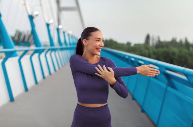 Mutlu kadın kollarını açıyor, antrenmandan önce ısınıyor, koşmaya hazırlanıyor, köprüde duruyor, yana bakıyor ve gülümsüyor.