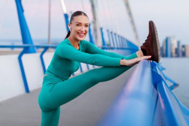 Heyecanlı sportif kadın antrenmandan önce ısınıyor, bacaklarını açıyor köprü korkuluklarına dayanıyor, koşuya hazırlanıyor.