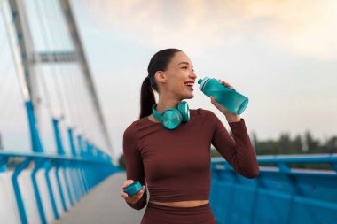 Koşu ve içme suyuna ara veren mutlu bayan, dışarıda antrenman yaparken kendini yenilemek için şişeyi tutuyor.