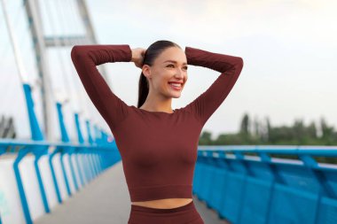 Mutlu sportif kadın ısınıyor, koşmadan önce kollarını esnetiyor, köprüde duruyor ve gülümsüyor.