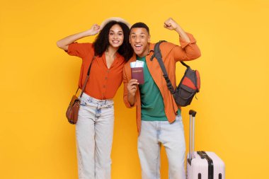Bavulları ve biletleri olan neşeli Afro-Amerikan çift, sarı stüdyo arka planında poz vererek zafer jestleri yapıyorlar ve yumruklarını sallıyorlar.