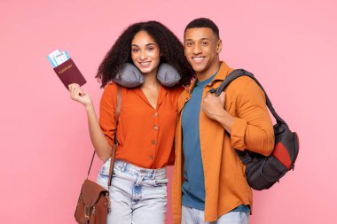 Biletleri ve sırt çantasıyla mutlu Latin çift, tatile hazır, pembe stüdyo arka planında duran, boyun yastığı takan kadın