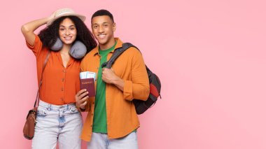 Pasaportlu ve sırt çantalı mutlu siyah çift pembe stüdyo arka planında poz veriyor, kameraya gülümsüyor, gezintiye hazır, boş alanı olan panorama