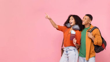 Mutlu Afrika kökenli Amerikalı çift tatile çıkıyor, pasaportlu kadın biletleri tutuyor ve boş yeri işaret ediyor, erkek sırt çantası taşıyor, harika bir seyahat teklifi sunuyor, pembe arka plan.