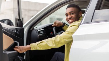 Pozitif siyah adam arabadan çıkıyor, otomobil kapısını açıyor ve gülümsüyor, içeri giriyor. Otomatik test sürüşü, satın alma, kiralama kavramı