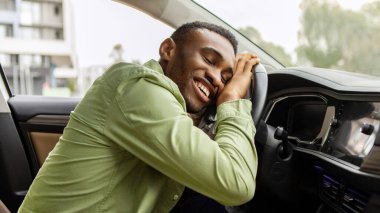 Şöför koltuğunda oturan, yeni arabasının direksiyonuna sarılan ve gülümseyen, rüyalarının tadını çıkaran keyifli siyahi adam.