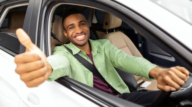 Mutlu Afrikalı Amerikalı adam lüks araba kullanıyor, başparmağını kaldırıyor ve gülümsüyor, aracın içinde oturuyor, rahat otomobillerin tadını çıkarıyor.