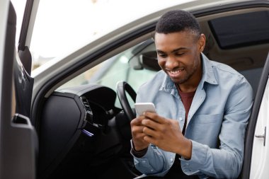 Cep telefonu kullanan mutlu Afrikalı Amerikalı adam, açık kapılı bir arabada oturuyor, GPS navigasyon uygulaması kullanıyor veya mesaj atıyor ve gülümsüyor.