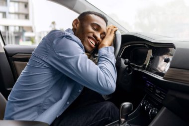 Şöför koltuğunda oturan, yeni arabasının direksiyonuna sarılan ve gülümseyen, rüyalarının tadını çıkaran keyifli siyahi adam.