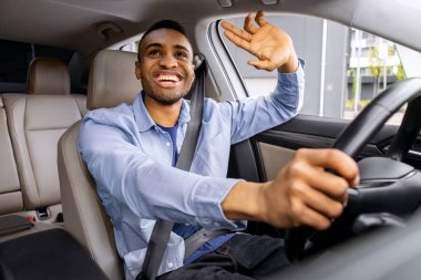 Pozitif siyah adam otomobil sürüyor ve el işareti yapıyor, birini selamlıyor, sürücü koltuğunda oturuyor ve direksiyonu tutuyor, araba sürüyor ve selam veriyor.