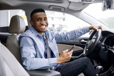 Modern arabanın içinde oturmuş kameraya gülümserken akıllı telefon kullanan pozitif siyah adam. GPS navigatör uygulaması ya da mesaj kullanan bir adam