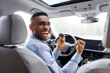 Yeni arabasında oturan, elinde araba anahtarı olan ve EVET işareti yapan, modern otomobilin galeride satın alınmasını kutlayan, dikiz manzaralı, aşırı neşeli siyahi bir adam.