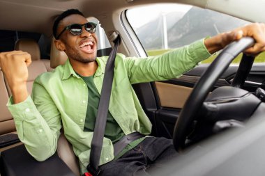 Güneş gözlüklü Afrikalı Amerikalı adam arabada dans ediyor, müzik dinliyor ve sürücü koltuğunda otururken şarkı söylüyor.