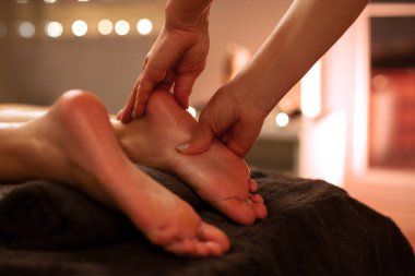 Profesyonel masör, genç bayan müşteriye ayak masajı yapıyor. Ayaklarını bastırıyor. Enerji akış noktalarını nazikçe etkiliyor.