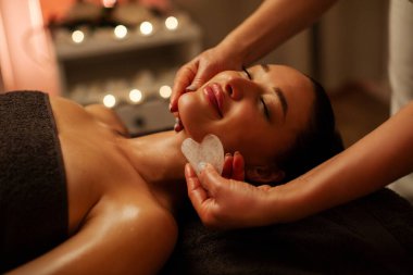 Genç Avrupalı bir kadın güzellik salonunda gua sha aracıyla profesyonel yüz masajı yaptırıyor. Gözleri kapalı yatan ve gülümseyen bir kadın.
