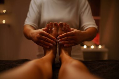 Genç bir kadının, mumlarla süslenmiş koyu renk spa salonundaki profesyonel masörden ayak masajı yaptırdığı pov görüntüsü.