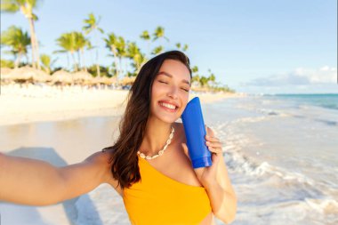 Güneş kremi tutan genç bir kadın, güneş ışınlarına karşı koruma öneriyor, sahilde selfie çekiyor. Yaz tatili