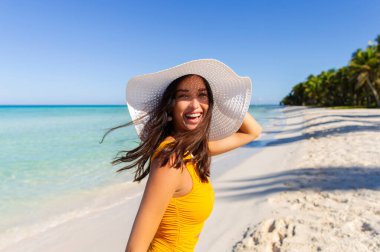 Sahilde hasır şapka takan, arka planında turkuaz okyanus olan güzel, mutlu bir bayan kameraya bakıyor ve gülüyor, tropikal tatilin tadını çıkarıyor.