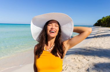 Hasır şapka takmış, gözleri kapalı gülümseyen, açık mavi okyanusla sahilde duran heyecanlı bir kadın.