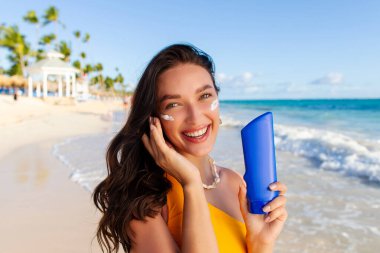 Güzel bayan bronzlaşmış yüzüne güneş kremi sürüyor ve tasarım için boş şişeyi gösteriyor, kumsalda duruyor ve kameraya gülümsüyor.