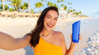 Güneş kremi şişesi gösteren mutlu bir kadın, okyanus kıyısında sahilde yürüyor, selfie çekiyor ve kameraya gülümsüyor.