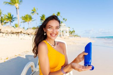 Mutlu genç bir kadın yüzüne güneş kremi sürüyor, plajda güneşlenirken güneşleniyor ve kameraya gülümsüyor.