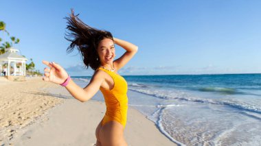 Mayo giymiş neşeli bir kadın mutlu bir şekilde kameraya bakıyor güneşli bir günde okyanus kıyısında koşuyor ve poz veriyor.