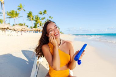 Kumsalda güneşlenip güneş kremi süren, güneş kremi kullanan ve gülümseyen mutlu bir kadın.