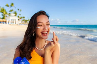 Neşeli bronzlaşmış kadın yüzüne güneş kremi sürüyor, mayo giyiyor, tropik okyanus kıyısındaki kumsalda duruyor. Güneş koruma cildi konsepti