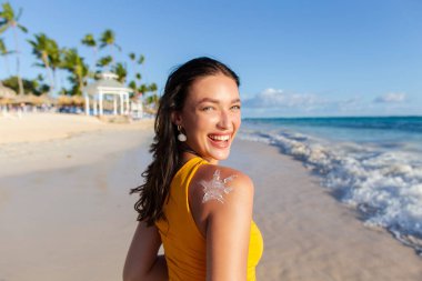 Omzunda güneş kremi olan genç bir kadın. Sahilde poz veriyor. Arka planda palmiye ağaçları ve okyanus var.