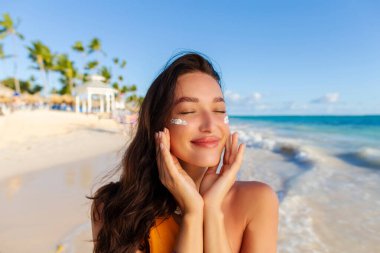 Yüzünde güneş kremi olan güzel bir kadın sahilde poz veriyor. Güneş koruması için cildine nemlendirici losyon sürüyor.