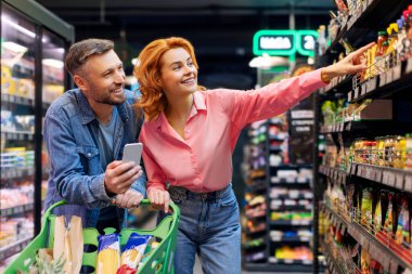 Mutlu eşler seçiyor, market alışverişi sırasında gıda ürünleri alıyor, modern süpermarkette rafın yanında duruyor, müşteriler esasları mağazada satın alıyor.