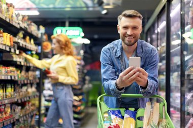Pozitif adam karısıyla market alışverişi yapıyor, erkek akıllı telefon kullanıyor süpermarkette arabaya yaslanıyor, eşler birlikte yemek alıyor.