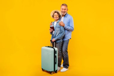 Seyahat biletleri ve bavulları olan mutlu yaşlı eşler sarı arka planda poz veriyor, kameraya gülümsüyor, tatil gezisine hazır, emekliliğin tadını çıkarıyor.