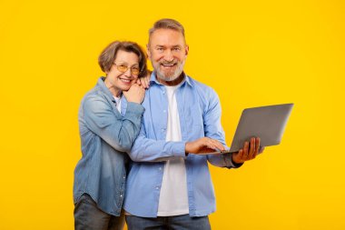 Üst düzey çiftler modern dizüstü bilgisayarda internette gezinmeyi seviyorlar, sarı arka planda bilgisayar başında duruyorlar, emekliliklerinde teknolojinin kolaylığını gösteriyorlar.