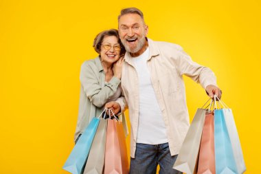 Mutlu, yaşlı çift kucaklaşırken, satın aldıklarıyla poz verirken ve sarı arka planda kameraya gülümserken. Büyük mevsimlik satışlar